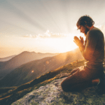 Person kneeling before natural scene with hands in prayer position and sun rising, in the mountains.